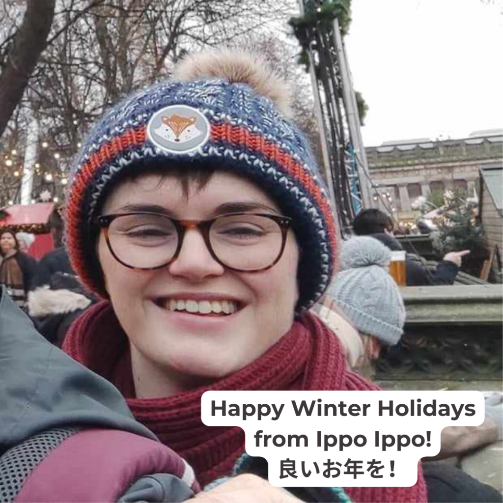 Elly grins at the camera, wearing a blue hat with a fluffy bobble at the back and fox cartoon image at the front. They're at the Edinburgh Christmas Market. Text reads: "Happy Winter Holidays from Ippo Ippo! 良いお年を！"