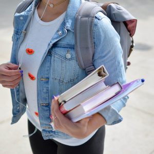 A student in a denim jacket carries textbooks in one arm. They're wearing a backpack and listening to something through a set of earphones.